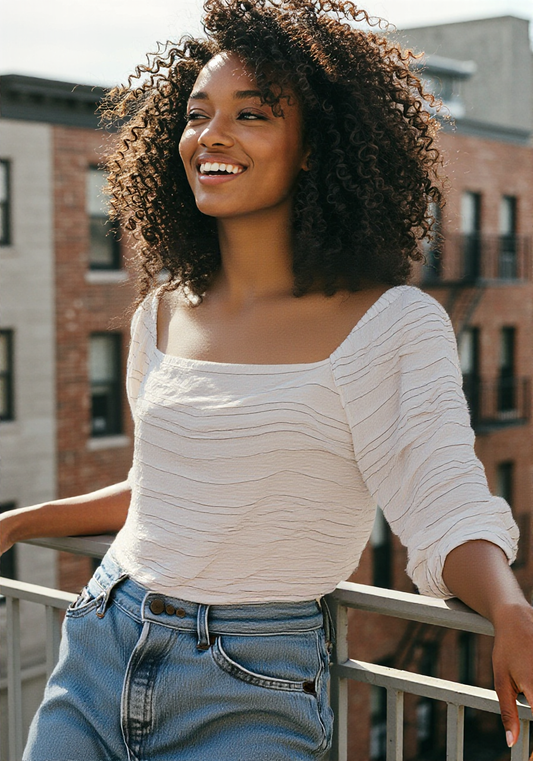 Woman standing on a balcony wearing a white top and blue jeans, with a cityscape background.